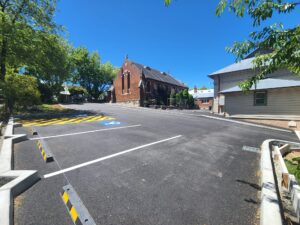 A newly paved, empty parking lot with a designated handicap space marked in blue and yellow. On the left, there are green trees providing shade. In the background, two brick and wooden buildings under a clear blue sky.