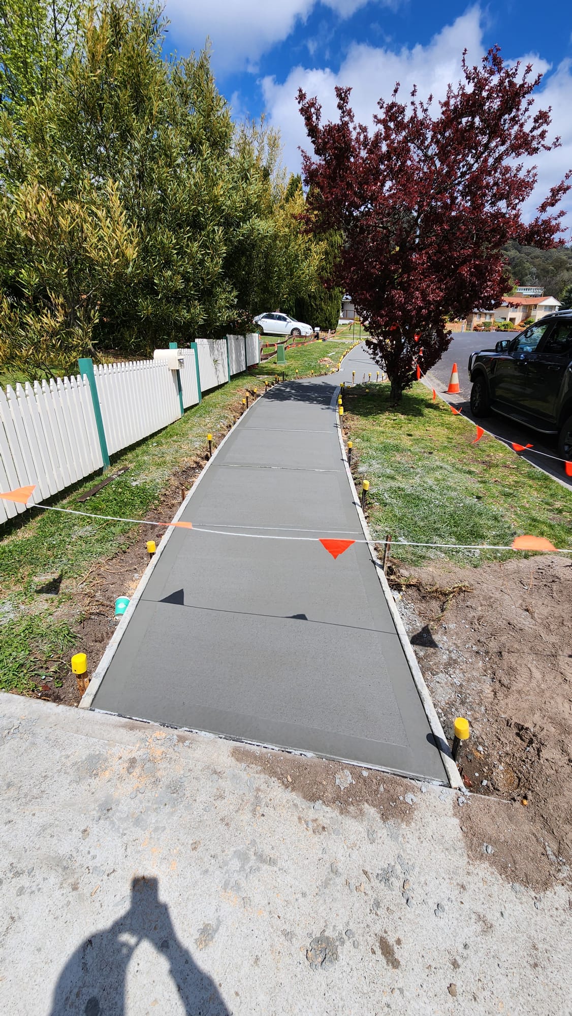 A freshly paved concrete sidewalk marked with orange cones and tape curves through a grassy area. Trees with green and red leaves line the path, and a white picket fence is on the left. A black vehicle is parked on the road next to the sidewalk.