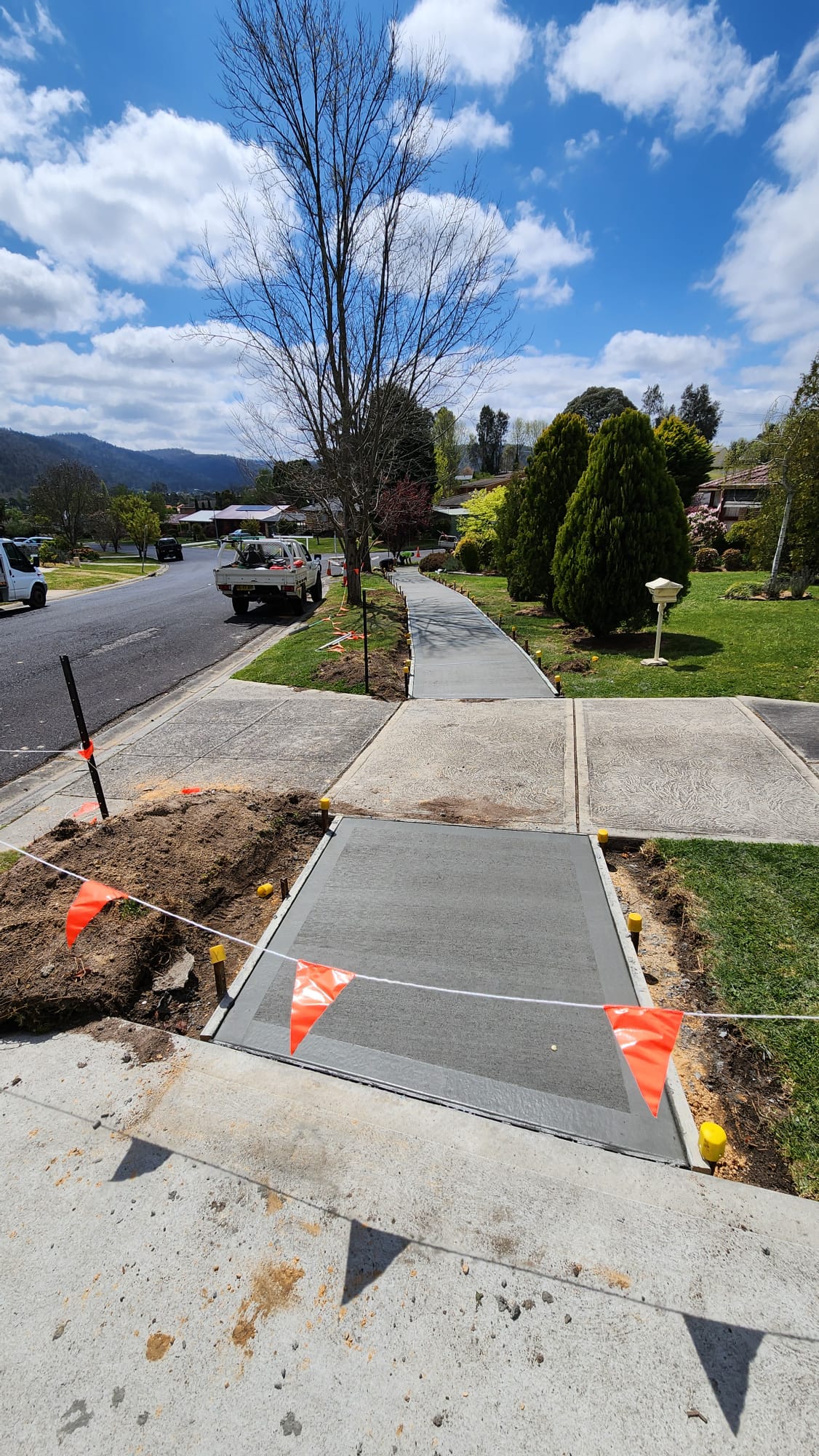 Freshly laid concrete path sectioned off with orange flags, running alongside a residential street with a parked white truck. Leafless tree and green shrubs border the sidewalk under a bright, partly cloudy sky.