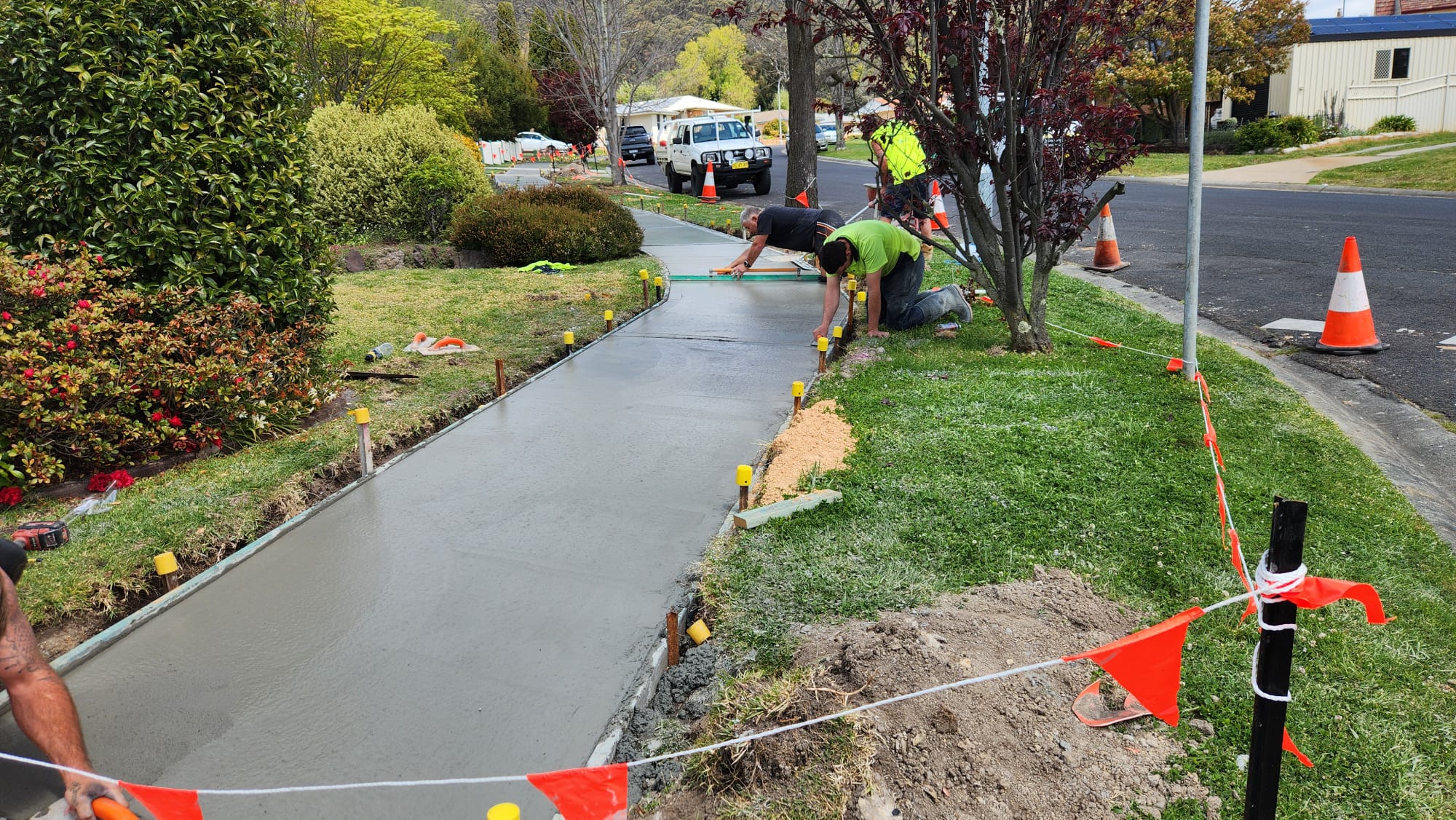 Workers are smoothing wet concrete on a section of sidewalk beside a road. Traffic cones and safety tape mark the area, with trees and shrubs in the background. A truck is parked nearby, and various tools and equipment are visible.