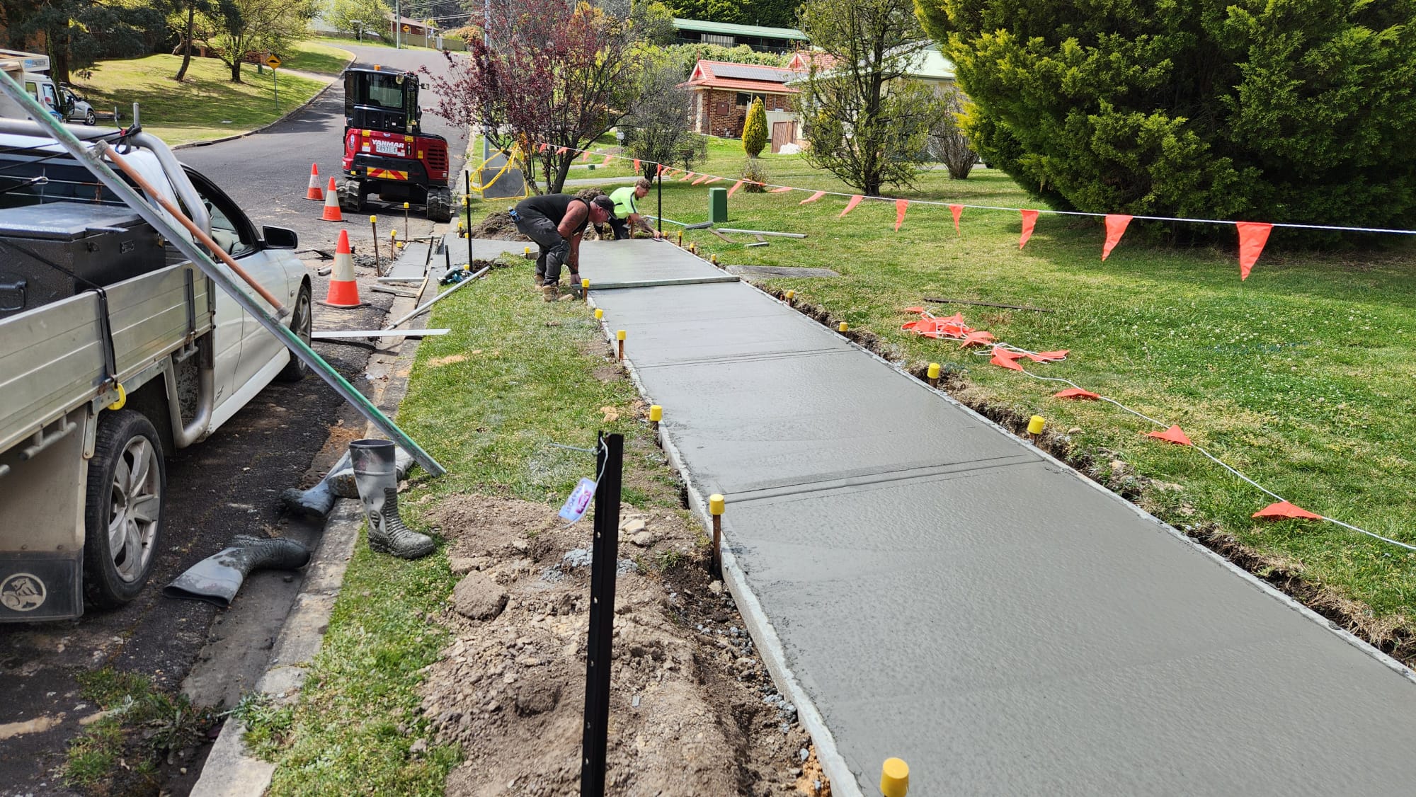 Workers are smoothing wet concrete on a newly laid sidewalk in a suburban area. Traffic cones and orange flags line the path. A truck is parked nearby on the road, and grass surrounds the work site.