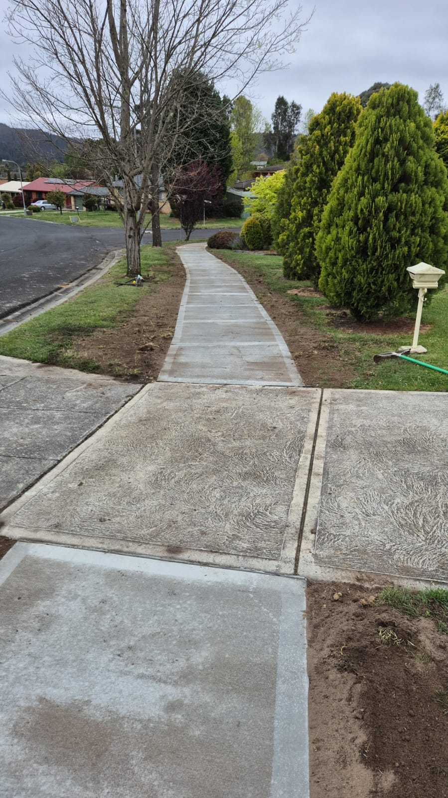 A freshly paved concrete path leads through a suburban neighborhood, bordered by trimmed grass and bushes, with trees lining one side. Overcast sky above and a street visible to the left.