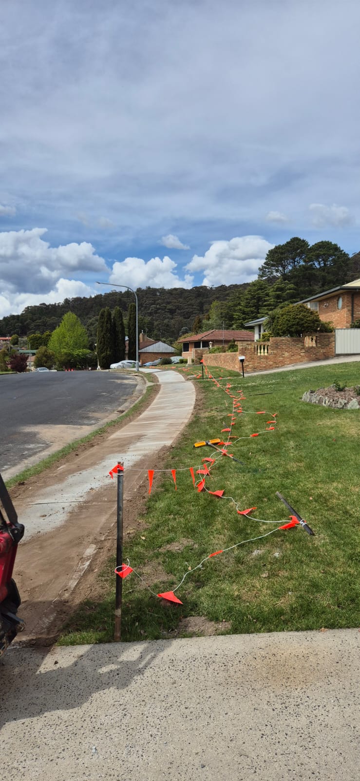 A newly constructed sidewalk curves along a suburban street with green grass beside it. Red flags are attached to a wire that marks the boundary of the grass. Houses and trees are visible under a partly cloudy sky in the background.