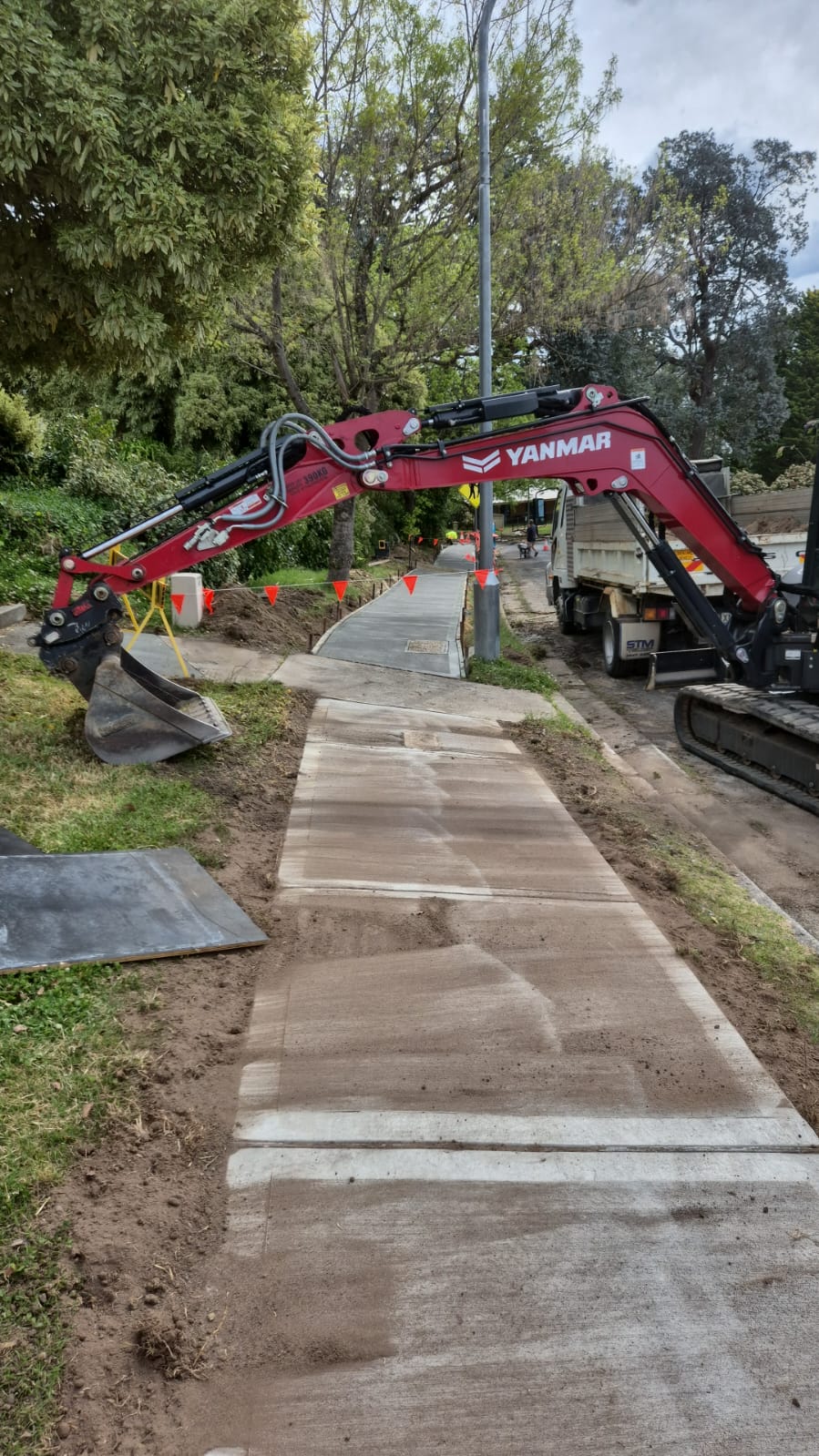 A construction site with a red excavator digging near a sidewalk. The path is partially covered with sand, and red warning flags are strung along the sides. Trees and greenery are visible in the background.