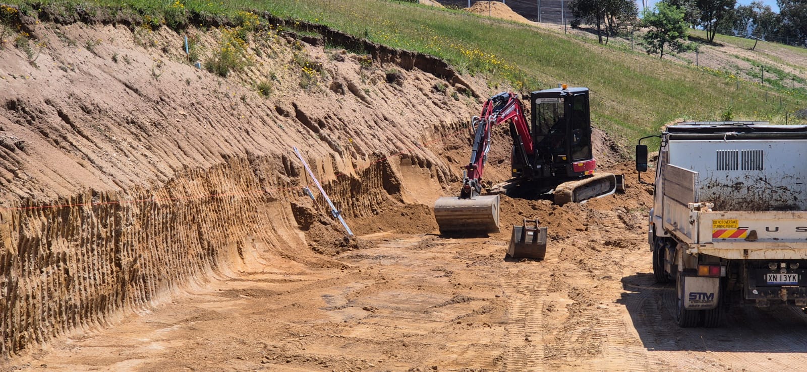 A construction site with an excavator digging earth on a sloped, grassy hill. A dump truck is parked nearby, and soil is piled around. The sky is clear and sunny, and trees are visible in the background.