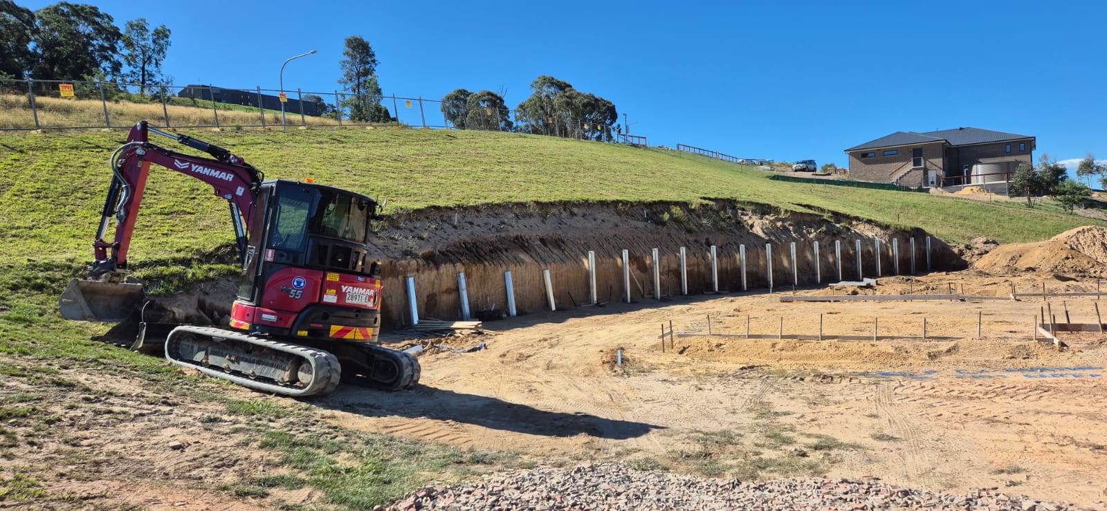 A red excavator works on a grassy slope, where a retaining wall with evenly spaced vertical concrete posts is under construction. Nearby, foundations and soil preparations are visible under a clear blue sky.