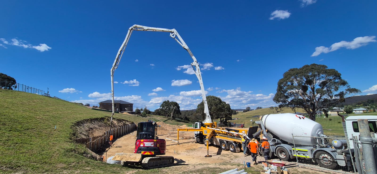 Construction site with workers in orange vests operating machinery, including a cement truck and a pump truck with an extended arm. A small excavator is also present. The background features grassy hills, trees, and a house under a blue sky.