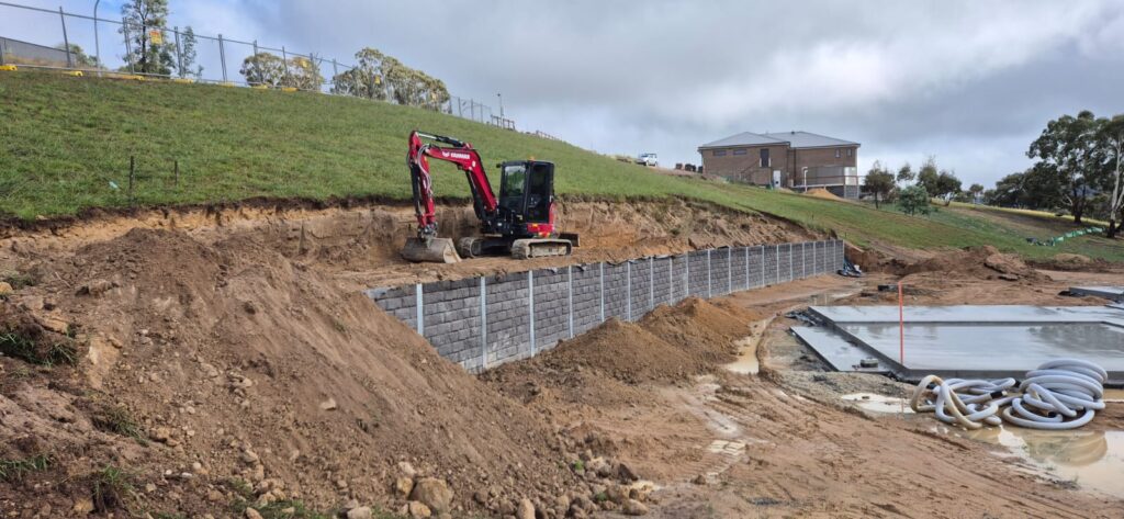 Construction site with a red excavator beside a partially built retaining wall on a muddy slope. The ground is uneven and wet, with a flat concrete area nearby. A house and trees are in the background under a cloudy sky.