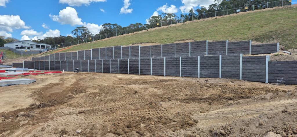 A construction site features a large terraced retaining wall built from dark blocks, set against a grassy hillside. The sky above is partly cloudy, and the area in front of the wall is covered in dirt and tire tracks.