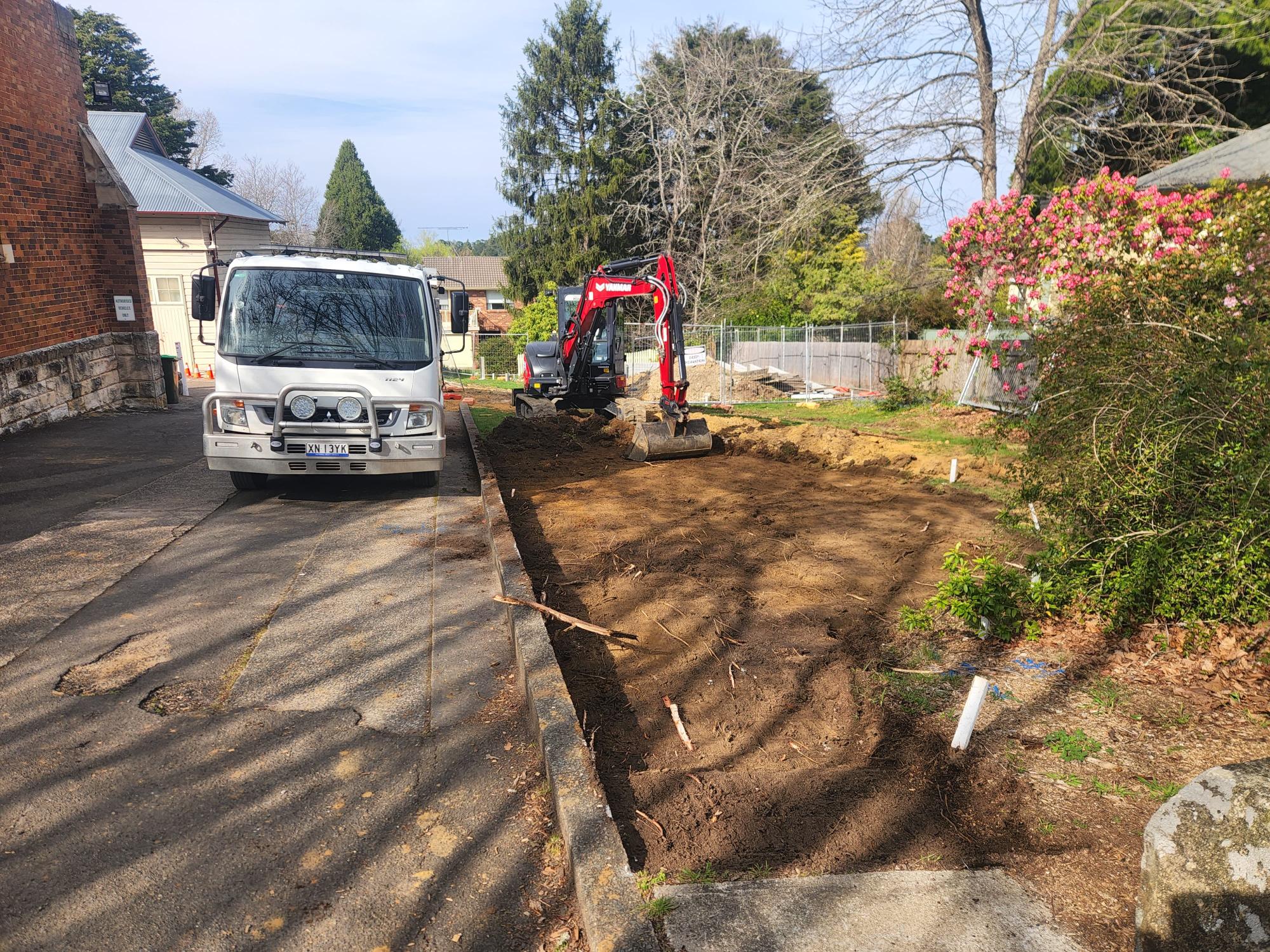 A small excavator is digging soil in a yard next to a brick building. A white truck is parked on the left. There are residential houses in the background and blooming flowers and trees nearby. The scene is outdoors on a sunny day.