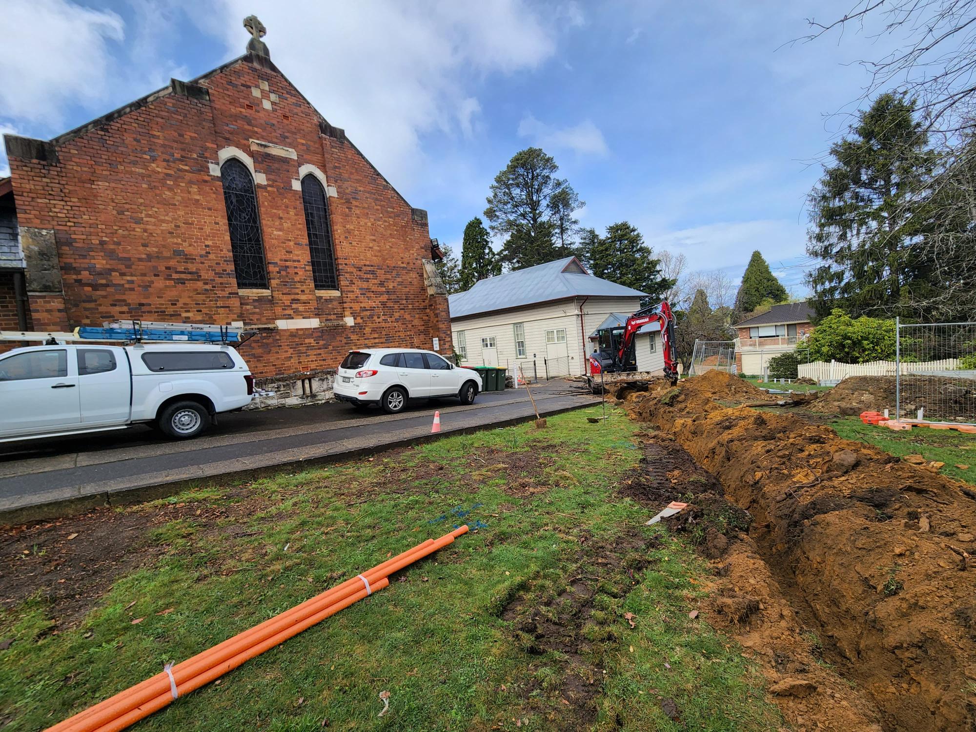 A construction scene near a brick building, with a trench dug into the grass. Orange pipes lie on the ground. A white pickup truck and an SUV are parked on the street. Trees and a white house are visible in the background.