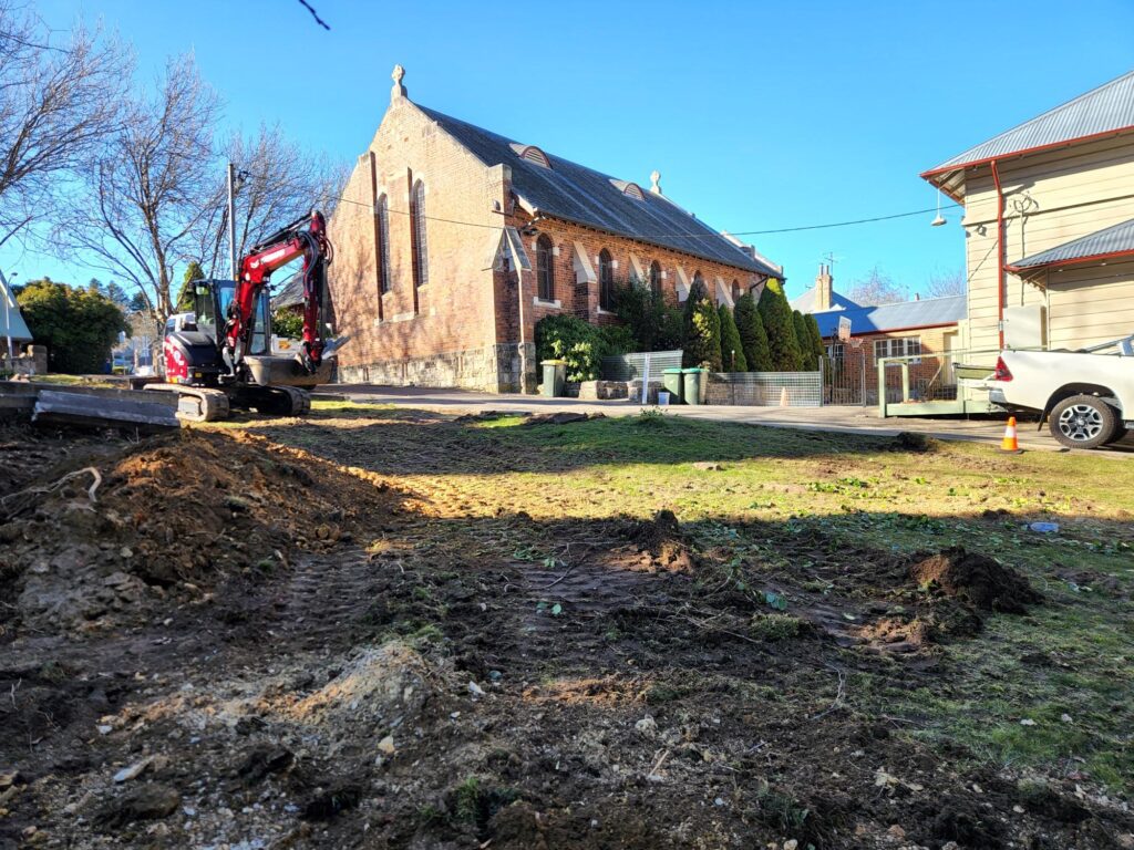 A small excavator is parked on a patch of dirt near a street. A historic brick building with arched windows is in the background, and a white utility vehicle is on the right. The scene is under a clear blue sky.