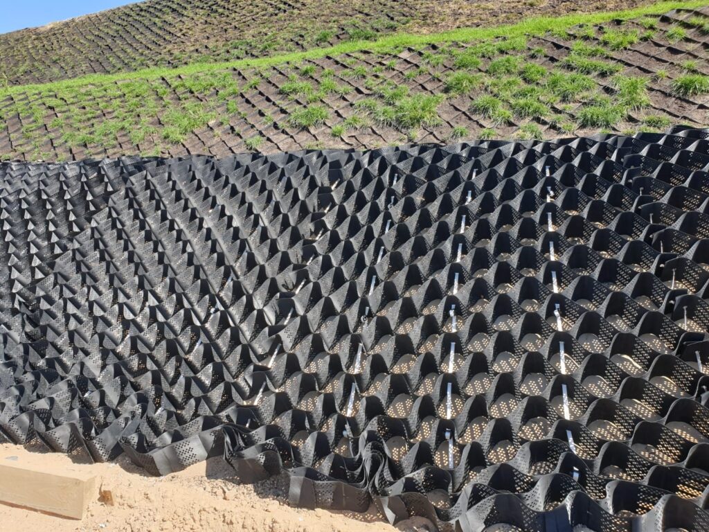 A grid-like erosion control geocell is laid out on a sloped surface, partially covered with soil and grass. The black plastic structure stabilizes the earth, aiding in landscape management and preventing soil erosion.