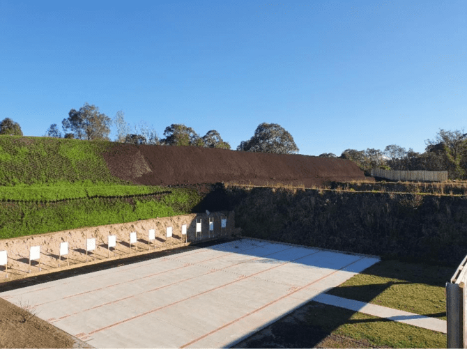 Outdoor shooting range with a concrete firing area and multiple target stands set against an earth berm. The background features grass and trees under a clear blue sky.