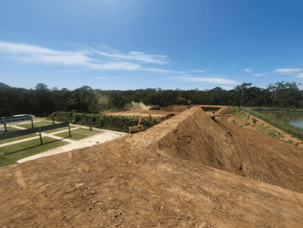 Construction site with bulldozers moving earth on a large dirt mound. Trees and a body of water are in the background under a clear blue sky. Dust is visible rising from the machinery in operation.