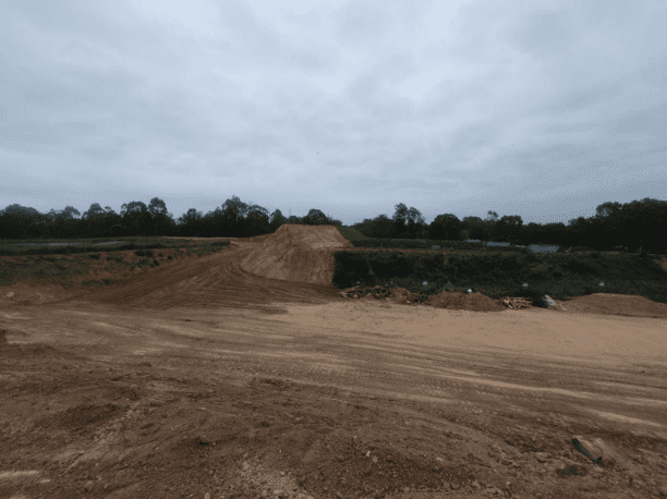 A barren construction site with dirt mounds under a cloudy sky. The foreground shows a wide, graded area of earth, and the background has trees lining the horizon.