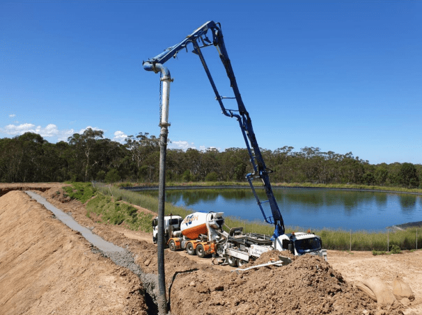 A construction site with a concrete pump truck pouring concrete into a trench next to a pond. The truck's boom arm extends over the trench, surrounded by dirt mounds. Trees and a clear blue sky are in the background.