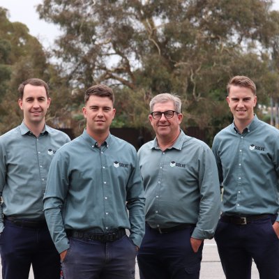 Four men wearing matching blue button-up shirts with a logo stand outdoors. They are posing together in front of trees and a fenced area. The sky is overcast.